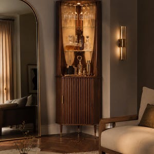 May include: A wooden corner bar cabinet with glass shelves, holding glassware and liquor bottles. The cabinet has a ribbed door and brass accents. A gold-framed mirror and a wall sconce are also visible.