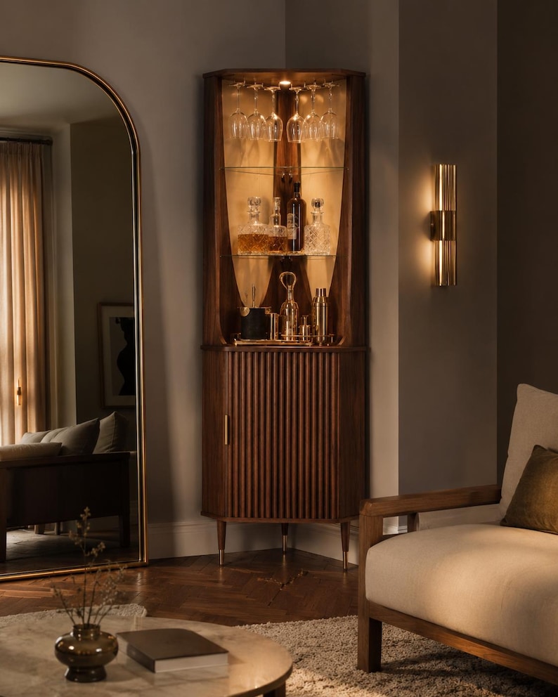 May include: A wooden corner bar cabinet with glass shelves, holding glassware and liquor bottles. The cabinet has a ribbed door and brass accents. A gold-framed mirror and a wall sconce are also visible.