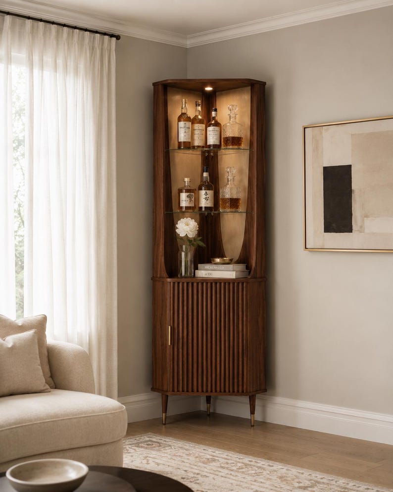 May include: A dark wood corner cabinet with glass shelves displaying liquor bottles and decanters. The cabinet has a ribbed door and brass-colored feet. A vase of white flowers sits on a shelf. The cabinet is in a well-lit room.