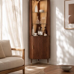 May include: A wooden corner cabinet with a warm brown finish. The cabinet features glass shelves displaying bottles and glassware. The lower section has a ribbed door with a gold handle. The cabinet is set in a well-lit room with a neutral color palette.