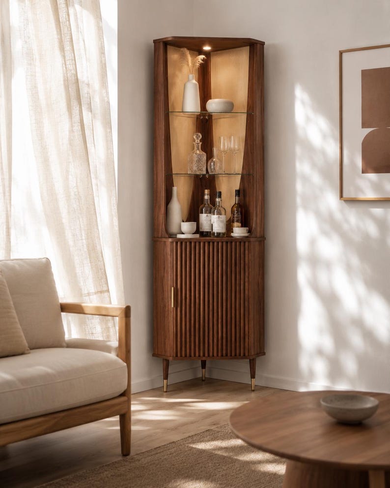 May include: A wooden corner cabinet with a warm brown finish. The cabinet features glass shelves displaying bottles and glassware. The lower section has a ribbed door with a gold handle. The cabinet is set in a well-lit room with a neutral color palette.