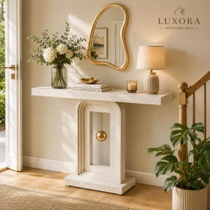 May include: A white marble console table with a gold accent mirror and a lamp. The table has a unique arched design with a hanging gold sphere. A vase of flowers and decorative items sit on the table. The text "Luxora - Interiors Nest" is visible.