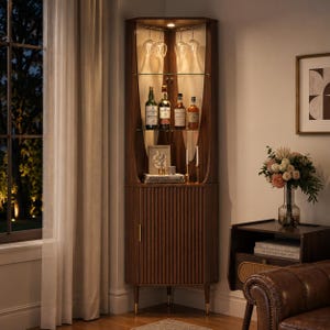 May include: A dark wood corner bar cabinet with glass shelves, holding bottles and glasses. The cabinet has a ribbed door and is lit from within. The cabinet is in a well-lit room with a window and a leather sofa.