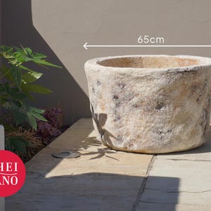 Op de afbeelding: Een grote, cilindervormige stenen plantenbak met een verweerd, gebroken wit en beige gestructureerd oppervlak. De plantenbak heeft een brede, open bovenkant en meet 65 cm in diameter en 40 cm in hoogte, aangegeven door de witte maatlijnen.