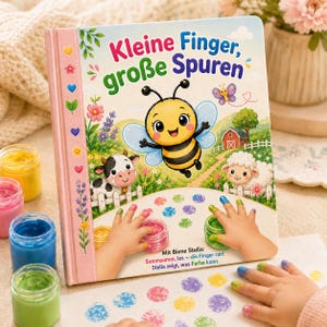 May include: A children's book titled "Kleine Finger, große Spuren" with a bee illustration. The book is open, showing colorful finger paint and handprints. Jars of paint in yellow, pink, blue, and green are visible.