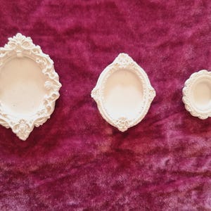 May include: Three white oval-shaped frames of varying sizes, each featuring intricate relief detailing along the edges. The frames are arranged horizontally, with the largest on the left, a medium-sized one in the centre, and the smallest on the right.