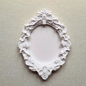 May include: White, ornate oval picture frame with intricate floral and bow details. The frame has a textured surface and a blank oval center. The frame is a decorative item.