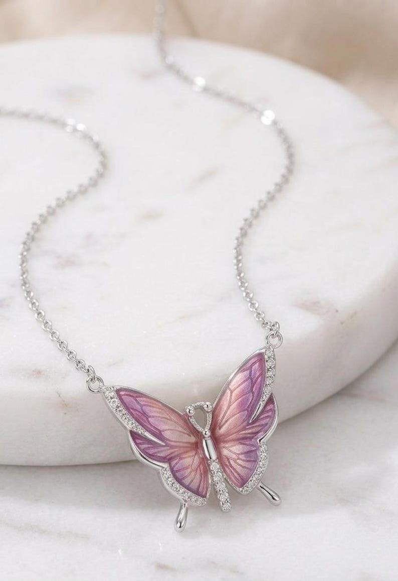 May include: A silver-toned butterfly pendant necklace. The butterfly has a gradient of pink and purple wings, outlined in silver, and accented with small, clear crystals. The necklace chain is delicate and silver.