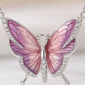 May include: A silver butterfly pendant necklace with a detailed design. The butterfly wings are a gradient of pink and purple, outlined with small, clear crystals. The pendant is a delicate piece of jewelry.