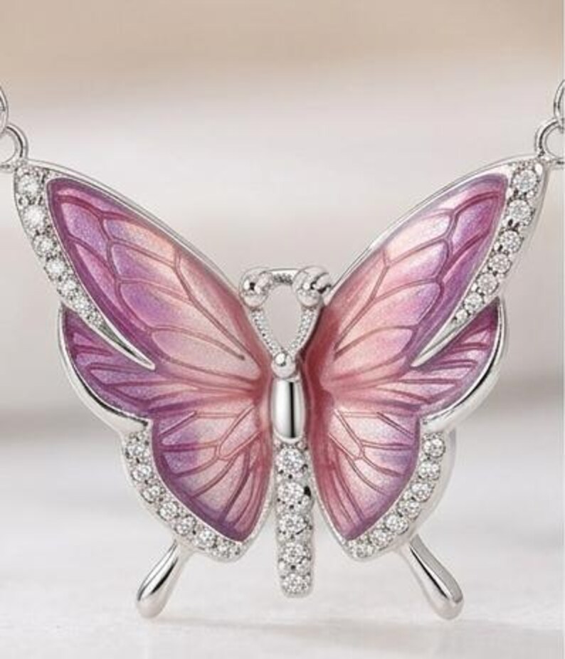 May include: A silver butterfly pendant necklace with a detailed design. The butterfly wings are a gradient of pink and purple, outlined with small, clear crystals. The pendant is a delicate piece of jewelry.