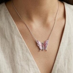 May include: A silver necklace featuring a pink butterfly pendant. The butterfly has detailed wings with a lighter pink hue and is adorned with small, sparkling accents. The necklace is worn against a neutral-colored top.