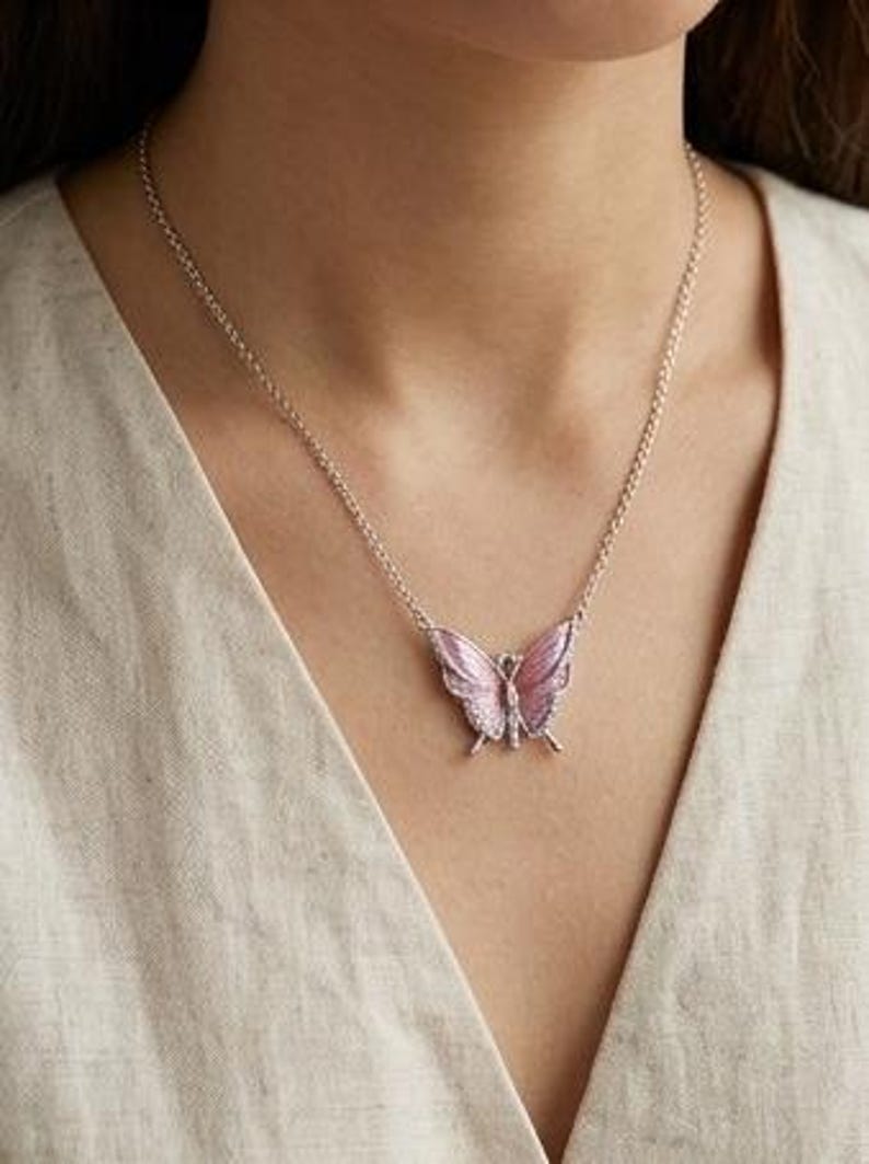 May include: A silver necklace featuring a pink butterfly pendant. The butterfly has detailed wings with a lighter pink hue and is adorned with small, sparkling accents. The necklace is worn against a neutral-colored top.