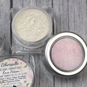 Könnte beinhalten: Drei kleine Gläser mit Schimmerstaub, beschriftet mit "Etherealle Pixie Gems Shimmer Dust - Love Potion". Die Gläser sind mit weißem, rosa und schimmerndem Schimmerstaub gefüllt.