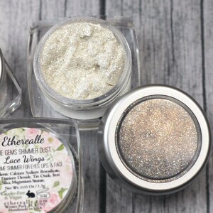 Puede incluir: Tres pequeños frascos de polvo brillante, etiquetados como "Etherealle Pixie Gems Shimmer Dust Lace Wings", sobre un fondo blanco. Los frascos están llenos de un polvo fino y brillante en tonos de blanco, dorado y plateado.