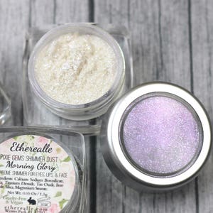 Peut inclure: Trois petits pots de poudre scintillante irisée, étiquetés "Etherealle Pixie Gems Shimmer Dust - Morning Glory Duochrome Shimmer for Eyes Lips & Face".