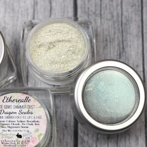 Peut inclure: Trois petits pots de poussière scintillante irisée, étiquetés "Etherealle Pixie Gems Shimmer Dust Dragon Scales Duochie Shimmer for Eyes Lips & Face".