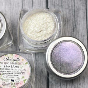 Peut inclure: Trois petits pots de poussière scintillante irisée, étiquetés "Etherealle Pixie Gems Shimmer Dust Dew Drops Doohickey Shimmer for Eyes Lips & Face".