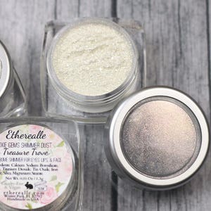 Peut inclure: Trois petits pots de poussière scintillante irisée, étiquetés "Etherealle Pixie Gems Shimmer Dust Treasure Trove". Les pots sont sur une surface en bois.
