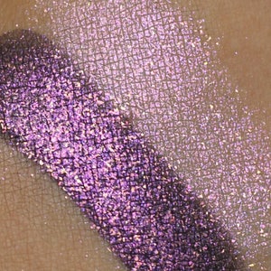 May include: A close-up of two eyeshadow swatches on skin. The left swatch is a deep purple with a shimmery finish. The right swatch is a light, iridescent white with a shimmery finish.