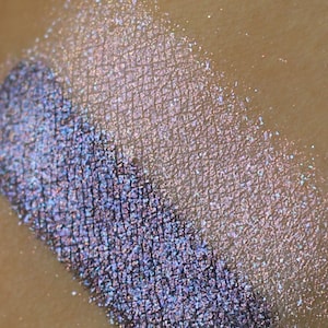 以下が含まれることがあります： キラキラと輝くパープルとホワイトのアイシャドウのクローズアップ。アイシャドウは細かいキラキラとした質感です。