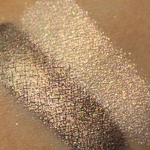 Puede incluir: Un primer plano de una muestra de sombra de ojos brillante en color dorado claro con un brillo iridiscente verde. La sombra de ojos está aplicada en la piel.