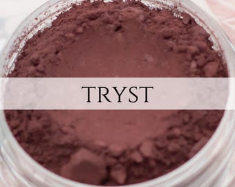 Op de afbeelding: Een close-up van een glazen pot gevuld met diep rood poeder met het label "TRYST".