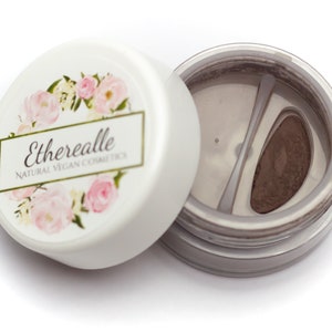 May include: A white round container with a floral design and the text "Etherealle Natural Vegan Cosmetics" on the lid. The container is open and shows a small, round, brown powder compact inside.