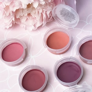 Puede incluir: Cinco pequeños recipientes redondos de rubor en crema en tonos rosa, melocotón y morado. Los recipientes son de plástico transparente con tapas blancas.