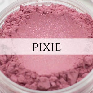 May include: A close-up of a pink blush powder with a shimmery finish. The powder is in a clear jar with the word "PIXIE" printed on a white label.