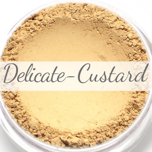 May include: A close-up view of a small, round container filled with a light yellow powder. The label on the container reads "Delicate-Custard".
