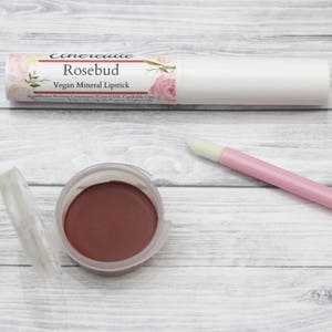 May include: A close-up of a rosebud-colored vegan mineral lipstick in a white tube with a floral design. The tube is labeled "Rosebud Vegan Mineral Lipstick". The lipstick is also shown in a small, clear container.