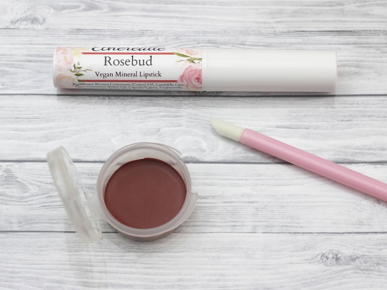 May include: A close-up of a rosebud-colored vegan mineral lipstick in a white tube with a floral design. The tube is labeled "Rosebud Vegan Mineral Lipstick". The lipstick is also shown in a small, clear container.