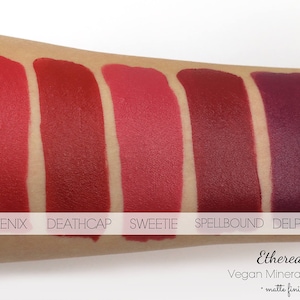 May include: Swatches of five matte finish vegan mineral lipsticks in shades of red and purple. The shades are named Phoenix, Deathcap, Sweetie, Spellbound, and Delphyne. The brand name Etherealle is also visible.
