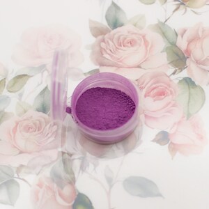 May include: A small, clear plastic container with a purple powder inside. The container is sitting on a white surface with a floral pattern.