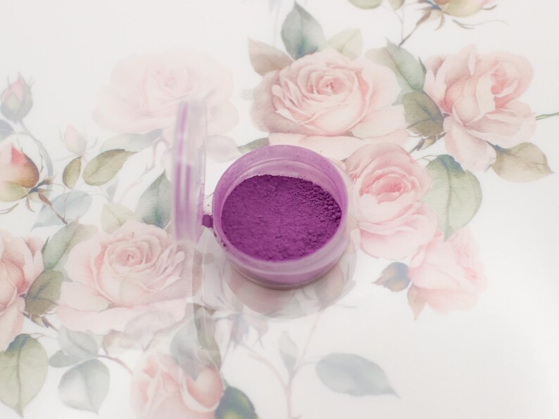 May include: A small, clear plastic container with a purple powder inside. The container is sitting on a white surface with a floral pattern.