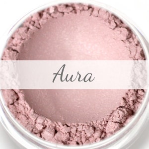 Puede incluir: Primer plano de un tarro lleno de polvo rosa claro. La palabra "Aura" está escrita en cursiva sobre una pancarta blanca. El polvo tiene una apariencia texturizada.