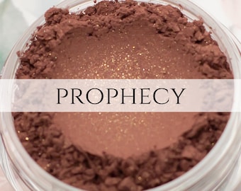 May include: A close-up of a brown eyeshadow with gold glitter. The label reads "PROPHECY".