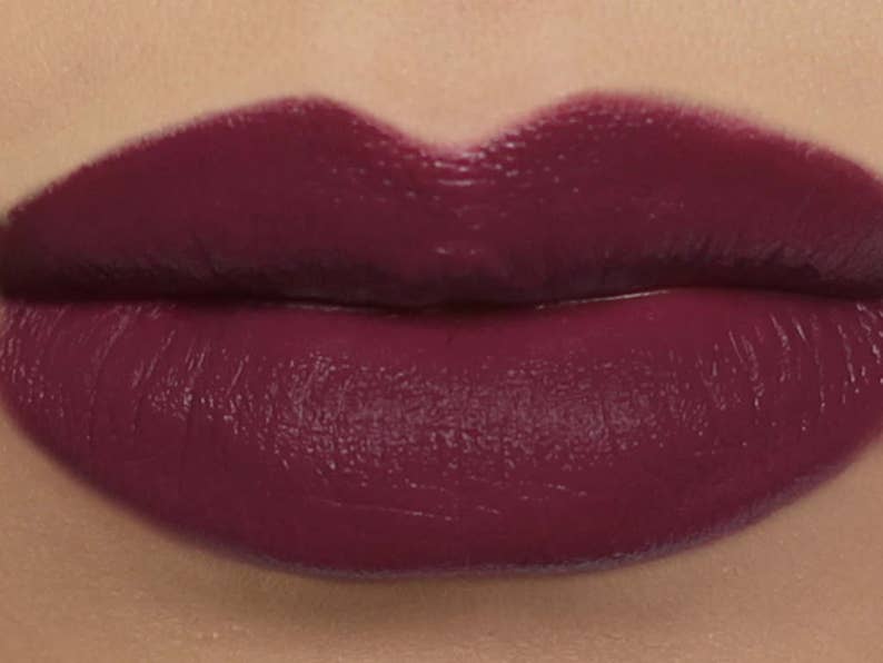 May include: Close-up of a person's lips, fully covered in a deep burgundy lipstick. The lipstick has a glossy finish, enhancing the rich color. The lips are well-defined, with a smooth and even application of the lipstick.