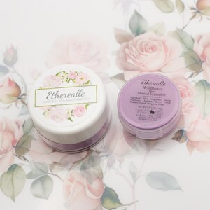May include: Two small jars of purple mineral eyeshadow with white lids and floral labels. The label on the left jar reads "Etherealle Natural Vegan Cosmetics". The label on the right jar reads "Etherealle Wildflower Bloom Mineral Eyeshadow".