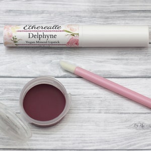 May include: A white lipstick tube and a small pot of dark red lipstick are displayed on a light wood surface. The lipstick tube has a floral label with the name "Delphyne." A pink applicator wand is also present.