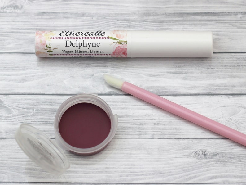 May include: A white lipstick tube and a small pot of dark red lipstick are displayed on a light wood surface. The lipstick tube has a floral label with the name "Delphyne." A pink applicator wand is also present.