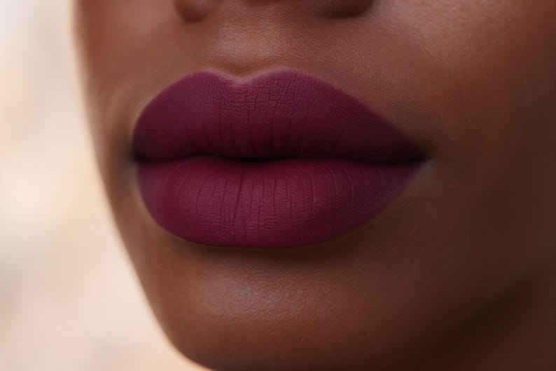 May include: Close-up of a person's lips painted with a matte, deep burgundy lipstick. The lipstick has a velvety texture and a rich, saturated color. The skin tone is dark, creating a striking contrast with the lip color.