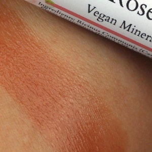 May include: Close-up of a brown lip balm tube with the text "Vegan Mineral Lip" and "Rosebud" on the label. The lip balm is applied to skin, showing a light brown color.