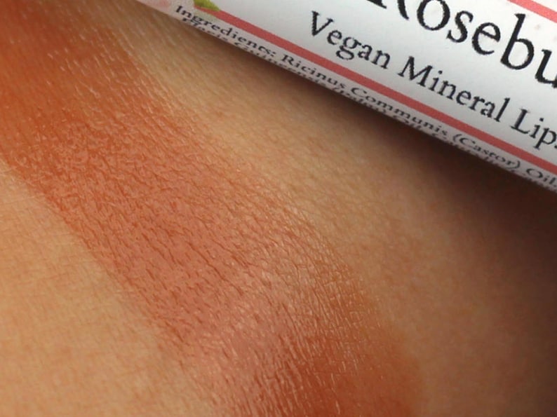 May include: Close-up of a brown lip balm tube with the text "Vegan Mineral Lip" and "Rosebud" on the label. The lip balm is applied to skin, showing a light brown color.