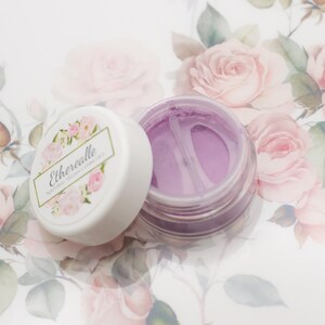 May include: A small, round jar of purple cosmetic powder with a white lid. The lid has the text "Etherealle Natural Vegan Cosmetics" printed on it. The jar is sitting on a white surface with a floral pattern.