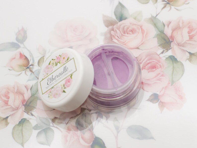 May include: A small, round jar of purple cosmetic powder with a white lid. The lid has the text "Etherealle Natural Vegan Cosmetics" printed on it. The jar is sitting on a white surface with a floral pattern.