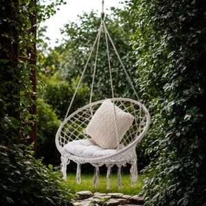 May include: A white macrame hanging chair with a cream cushion and pillow. The chair is suspended from a rope and hangs in a green garden. The chair has a woven net design and decorative fringe, perfect for relaxing.