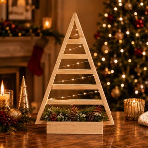 Op de afbeelding: Een driehoekig houten frame in de vorm van een kerstboom, voorzien van horizontale latten omwikkeld met dunne lichtsnoeren. De basis bevat een decoratief arrangement van groene dennentakken, kleine dennenappels en rode bessen.