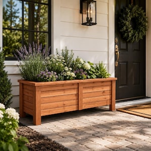 Puede incluir: Una jardinera rectangular de madera llena de varias plantas y flores. La caja es de madera de color marrón claro y se apoya sobre patas cortas. Las plantas incluyen lavanda, flores blancas y moradas. La jardinera está cerca de una puerta principal.