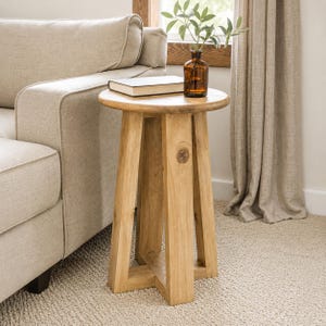 May include: A light-coloured wooden side table with a round top and four angled legs. A book and a small brown glass vase with greenery sit on top of the table. The table is next to a beige sofa.